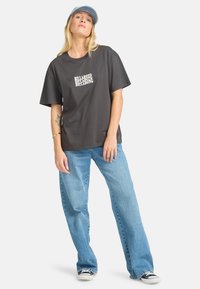 Camiseta oversize de color gris oscuro con el logo blanco de "BILLABONG", combinada con jeans rectos de color azul claro y zapatillas negras. El modelo lleva una gorra azul.