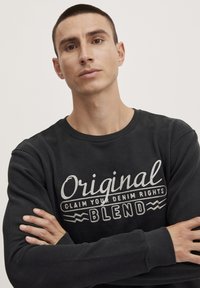 Černá mikina s kulatým výstřihem a dlouhými rukávy. Přední text bílou barvou, "Originální nárok na vaše džíny." Měkká textilie.