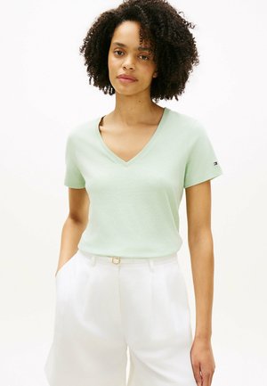 Femme aux cheveux bouclés portant un T-shirt à col en V vert clair et un short taille haute blanc, mains dans les poches, debout devant un fond uni.