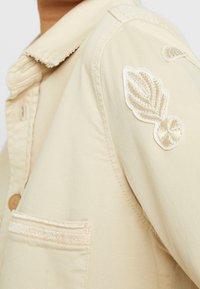Beige denimjacka med krage, prydd med en dekorativ broderad lövpatch på axeln och ett strukturerat horisontellt rand på fickan.