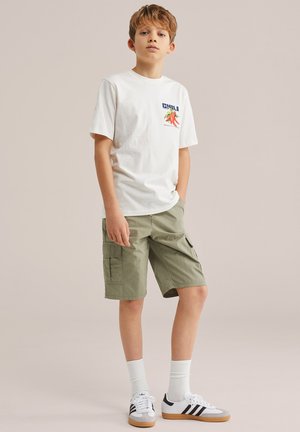Dreng stående med hænderne i lommerne iført hvid T-shirt med chili-grafik, grøn cargo-shorts, hvide sokker og hvide Adidas-sneakers.