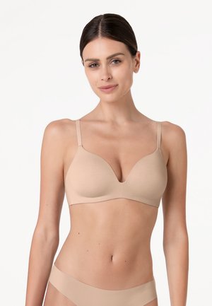 Donna con capelli scuri che indossa un reggiseno beige senza cuciture e biancheria intima coordinata, in piedi su uno sfondo bianco semplice.