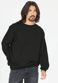 Musta crewneck-huppari, valmistettu pehmeästä materiaalista, jossa on ribbireunat hihoissa ja helmassa. Mallilla on päällään tummanharmaat farkut. Yksinkertainen muotoilu, ei kuvioita.