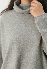 Donna che regola il colletto di un morbido maglione grigio chiaro a collo alto con maniche lunghe e polsini a coste.