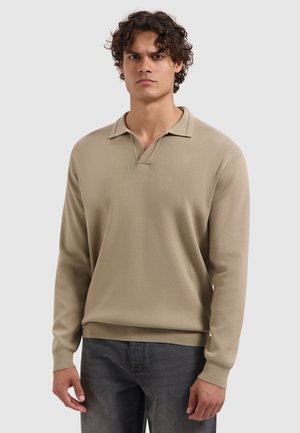 Man met krullend haar, gekleed in een beige lange mouwen gebreid polo sweater en donkergrijze jeans, staande tegen een effen lichte achtergrond.