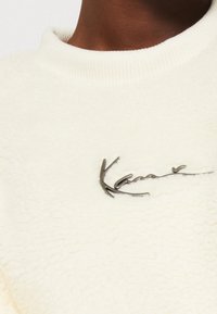 Krémově zbarvená fleecová mikina s texturovaným povrchem a kovovým logem uprostřed, s žebrovaným výstřihem.