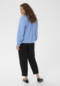 Bluză lungă de culoare albastru deschis cu manșete plisate, purtată cu pantaloni negri îngustați și pantofi negri fără toc, expusă dintr-o ușoară perspectivă din spate.