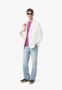 Blazer de lino blanco sobre una camiseta rosa, jeans azul claro y zapatillas oscuras. Los accesorios incluyen gafas de sol y un objeto doblado en la mano.