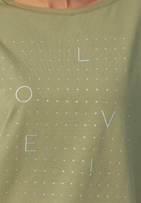 Olijfgroene t-shirt met een ronde halslijn. Bestaande uit zilveren folie tekst "LOVE" en ronde studs die in een ruitpatroon zijn gerangschikt. Zachte stofstructuur.