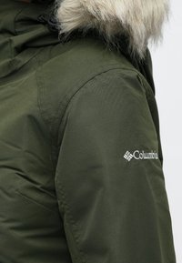 Zielona kurtka z kapturem i sztucznym futerkiem, o gładkiej fakturze, z kieszeniami po bokach i widocznym białym logo Columbia na rękawie.