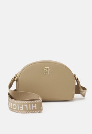 Beige halfronde crossbodytas met gouden logo aan de voorkant en verstelbare stoffen band bedrukt met witte tekst ""HILFIGER"".