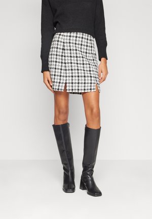 Woman wearing a black sweater, a white and black checkered skirt with front slits, and black knee-high heeled boots against a plain background.
