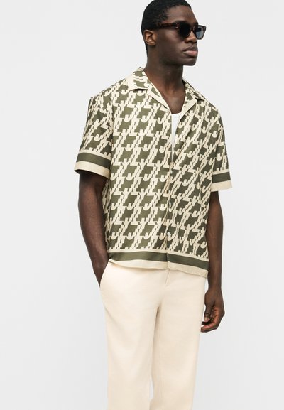 Άνδρας με σκούρα γυαλιά ηλίου, κοντομάνικο πουκάμισο σε μπεζ και ελαι πράσινο μοτίβο, μπεζ παντελόνι, στέκεται με το ένα χέρι στην τσέπη.