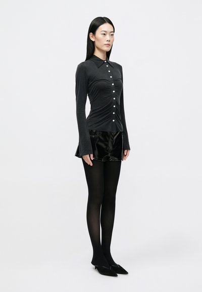 Long-sleeve black shirt with a collared neckline and button front, paired with shiny black shorts and opaque black tights, with pointed shoes.