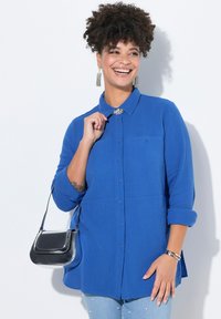 Camicia blu con bottoni, in un tessuto strutturato, maniche lunghe arrotolate fino ai gomiti, due tasche sul petto e abbinata a una borsa nera.