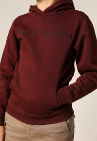 Sweat à capuche bordeaux en tissu doux, doté d'une poche avant, de poignets côtelés, et du texte "DELUXE ORIGINAL" en noir sur la poitrine.