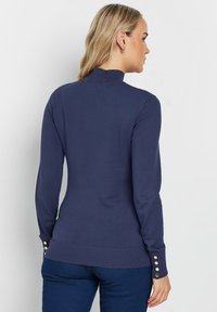 Navy-blauer Langarmpullover mit Stehkragen, geripptem Saum und Knopfdetails an den Ärmeln. Hergestellt aus einem glatten, leichten Stoff.