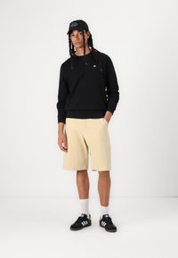Black sweatshirt, beige shorts, and black sneakers with white stripes. Black cap with white text. Simple, casual outfit.
