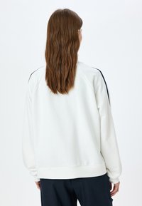 Sweatshirt blanc, doux, avec une coupe décontractée, présentant des manches raglan et des accents noirs sur les épaules. Texture lisse, ourlet côtelé.