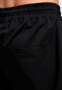 Pantalons en tissu noir avec une taille élastique légèrement froncée et une poche arrière unique, présentant une texture lisse et mate.