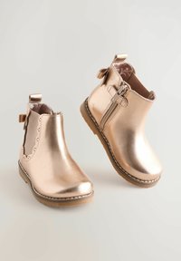 Stivaletti alla caviglia in rosa dorato metallico con una finitura liscia, bordo decorativo a onde, zip laterale e dettaglio a fiocco sul tallone.