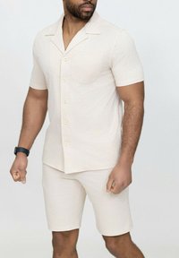 Chemise à manches courtes avec boutons et shorts assortis en beige clair avec un motif en chevrons. Le tissu semble doux, décontracté et avec une coupe ample.