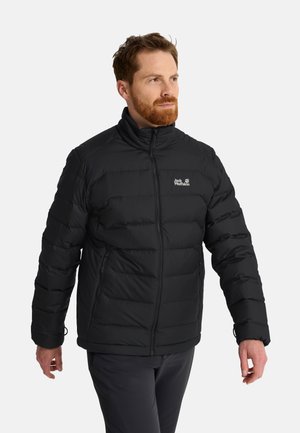 Mann mit rotem Bart, der eine schwarze gesteppte Jacke mit Reißverschluss und dunkle Hose trägt, blickt seitlich vor einem schlichten Hintergrund.