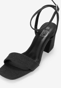 Sandal con tacco alto nero, con punta quadrata e tacco spesso. Presenta una singola fascia larga attraverso il davanti del piede e una sottile fascia alla caviglia, in tessuto strutturato.