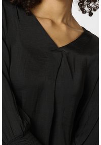 Femme portant un chemisier noir à col en V avec des manches bouffantes froncées et un tissu texturé, montrant son cou et ses cheveux bouclés.