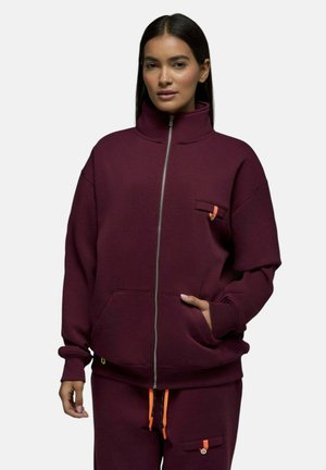 Femme portant une veste zippée et un pantalon bordeaux assortis avec des cordons orange et des poches boutonnées, debout devant un fond uni.