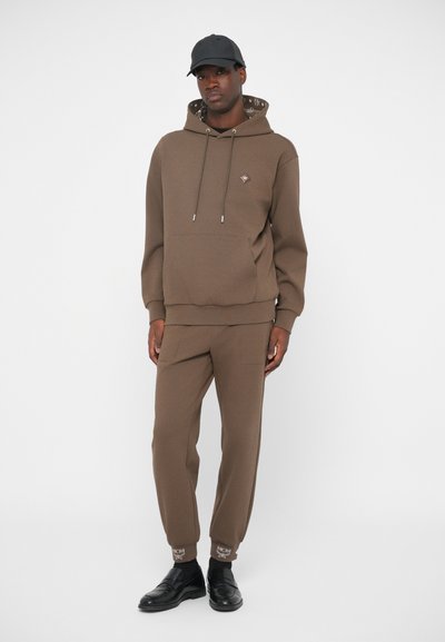 Brązowa bluza z kapturem i pasujące dresy wykonane z miękkiego materiału. Posiada naszywkę z logo, regulowane sznurki ściągające i prążkowane mankiety. Na nogach czarne buty.