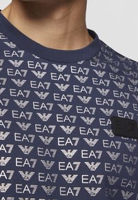 Camiseta de color navy con logotipos EA7 en blanco repetidos en un patrón geométrico; cuello redondo; etiqueta EA7 negra cosida en la parte inferior derecha.