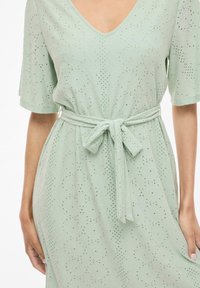 Ljusgrön klänning med V-ringning, korta ärmar och ett band i midjan. Tyget har ett strukturerat, prickigt mönster.