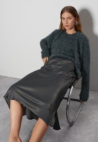 Texturierter türkiser Pullover mit lockerer Passform und langen Ärmeln, kombiniert mit einem glänzenden dunkelgrauen Satinrock, der sanft um die Beine fällt.