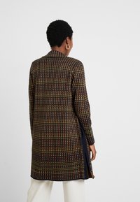 Tunique longue à motifs en tissu sombre avec un design à quadrillage multicolore. Présente des fentes sur les côtés et un col, avec une coupe ajustée.