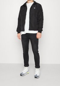 Czarna kurtka na zamek z wysokim kołnierzem i logo na piersi, zestawiona z białym t-shirtem, ciemnymi dżinsami i szarymi trampkami z neonowymi akcentami.