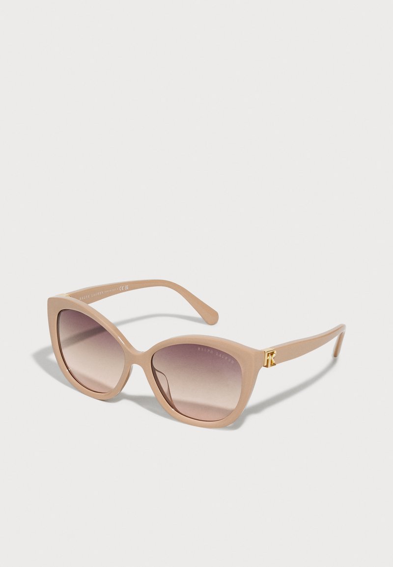 Óculos de sol estilo gato com uma armação suave em bege, lentes degradê e um detalhe em logo dourado na haste. Textura plástica suave.