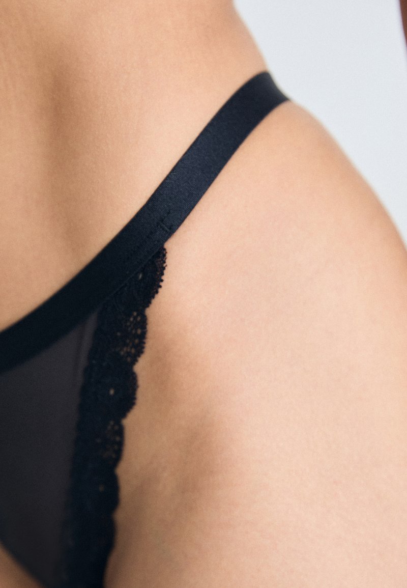 Zwarte thong met kantafwerking, gemaakt van gladde stof, met een strakke tailleband en gedetailleerde gekartelde randen aan de zijkanten.