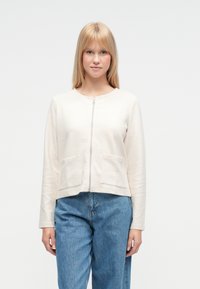 Hellbeige Reißverschlussjacke mit strukturierter Stoffoberfläche, abgerundetem Ausschnitt und zwei Vordertaschen. Kombiniert mit blauen, hoch taillierten Denim-Hosen.