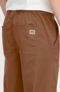 Brune bukser laget av bomull med elastisk midjebånd. Har to baklommer og et lite vevd merke på høyre lomme.