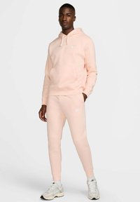 Sudadera Nike de color rosa claro con bolsillo frontal y logo, combinada con pantalones de chándal a juego que presentan el logo, fabricados en tejido suave, usados con zapatillas ligeras.