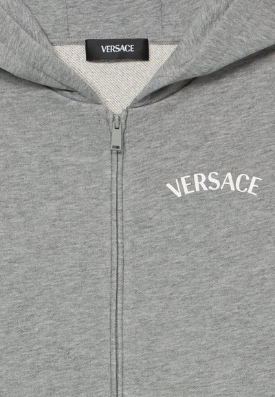 Graue Zip-Up-Hoodie mit weicher Textur, versehen mit einem schwarzen Etikett und dem weißen "VERSACE"-Logo über dem Reißverschluss in einem gewölbten Design.