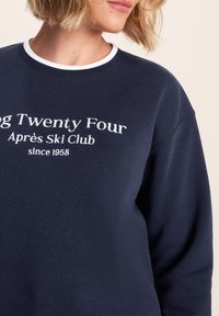 Navy blue sweatshirt with white ribbed collar, featuring embroidered text: "Twenty Four Apres Ski Club since 1958." Soft texture and loose fit.