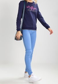 Sweatshirt marine avec logo "Hilfiger", jeans skinny bleu clair, baskets blanches et sac bandoulière teal, avec un design simple et une texture lisse.