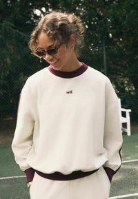 Biały sweatshirt z bordowym ściągaczem przy kołnierzu i mankietach, z haftem "self." w kolorze brązowym. Miękka tkanina, luźny krój, długie rękawy.