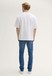 Camiseta de algodón blanca con mangas cortas, que presenta un ajuste relajado. Combinada con jeans de mezclilla azules y zapatillas blancas. Fondo liso.