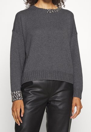 Femme portant un pull gris foncé avec des ornements en strass au niveau de l'encolure et des poignets, associé à un pantalon en cuir noir.