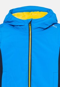 Blå jakke med hette i gul pels, hel lengde glidelås og svarte sidepaneler. Har en jevn tekstur og slitesterkt materiale.