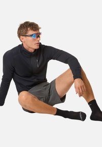 Sportbekleidung mit einer schwarzen Jacke mit Reißverschluss und grauen Shorts, ergänzt durch schwarze Socken und bunte Sonnenbrillen.