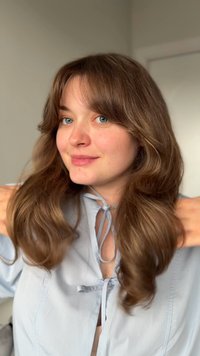 Ung kvinne med langt brunt hår og blå øyne som justerer en lys blå bluse med knyttede sløyfer i et lyst innendørs miljø.